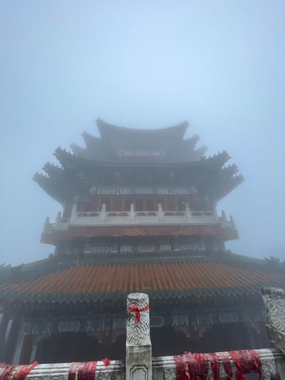 The image shows a portion of a traditional Chinese building shrouded in heavy fog. The building features a multi-tiered, ornate roof with upturned eaves. Details of the roof include orange-colored tiles and intricate carvings. The building's structure is visible, revealing red and white columns with decorative patterns. Red ribbons are tied around the columns. The fog obscures much of the building and surrounding environment. The photograph was taken in Yongding District, China.