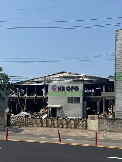 The image shows a two-story commercial building in Pyeongtaek-si, South Korea, that appears to have suffered significant fire damage. The roof and upper sections of the building are blackened and appear to be partially collapsed, with exposed structural elements. The windows on the upper floor are broken or missing, revealing the interior which is filled with debris and dark, charred materials. The lower floor has some intact windows and a door. A sign with the logo "KR OPO" in red and blue is visible on the front of the building, above a green stripe. In front of the building, there is a metal folding gate and a low brick wall. The area in front of the building and along the road is littered with debris, including what appears to be flattened metal sheets, wooden planks, and other unidentifiable objects. The road in the foreground has traffic cones and markings. The sky is clear and blue. There are no people clearly visible in the image.