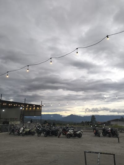 In Panteraya, Indonesia, a cloudy twilight scene unfolds over a gravel parking area in front of a building with an awning and string lights. A line of motorcycles is parked in the foreground, some with their headlights on. In the background, mountains are visible under a dramatic sky. The atmosphere suggests a remote, perhaps rural, setting.