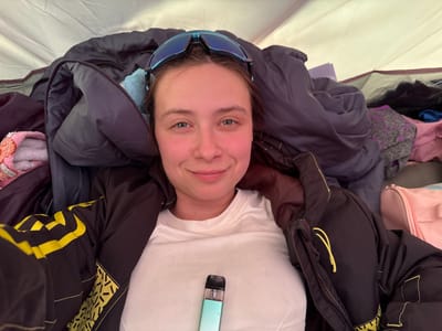 A woman is lying down, appearing to be inside a tent. She is wearing a white t-shirt and a black jacket with yellow accents. She has blue sunglasses resting on her forehead. A vape pen is positioned near her chest. Other clothing items and bags are scattered around her inside the tent. The location is unspecified and not identifiable from the image.