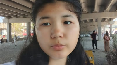The image captures a close-up selfie of a young East Asian woman. Her face fills the foreground, with her dark hair framing her face. Her expression is neutral, and her gaze is directed towards the camera. Behind her, the scene unfolds under an elevated structure, possibly a highway overpass or a large covered market.

In the background, several people are visible. To the left, a person is sitting on the ground. Further back, a person in a white garment with a head covering walks, carrying a bag. On the right side of the image, a man in a maroon shirt and dark pants stands with his back to the camera, facing away from the viewer. Next to him, a woman with a backpack is also standing. Further to the right, another person in a patterned green garment is visible. Cars are parked and driving in the distance, suggesting a public area.

The setting appears to be an outdoor urban environment. The presence of the elevated structure, buildings in the background, and parked cars indicates a city location. The lighting suggests it is daytime, likely sunny. There is some visible text on a yellow object in the lower right portion of the image, with the numbers "31" clearly legible. The overall impression is a candid moment captured in a busy public space.