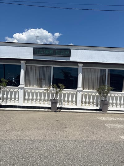The image presents a sunlit daytime scene in Psyrdzkha, Abkhazia, featuring the facade of a building next to an asphalt road. The building exhibits a classical architectural style, primarily white, with a balcony or terrace adorned with white, ornate balustrades and supported by elegant white columns. On this terrace, two grey cylindrical planters hold small, leafy green plants or shrubs.

The building's upper level features large, dark-tinted windows, some revealing light-colored curtains inside. Above these windows, a horizontal digital display with green, pixelated characters is mounted. The specific text is not clearly legible, but it appears to be an informational display, possibly showing numbers like a temperature reading or a scrolling message.

The upper portion of the image is dominated by a vibrant blue sky, hosting a single large, fluffy white cumulus cloud. Two thin, dark utility wires or cables stretch across the top-left of the sky. A small, multi-pronged antenna is visible on the building's roof, partly obscured by the cloud.

The foreground is occupied by an asphalt road, which shows faint, light-colored lines, possibly remnants of a pedestrian crossing. The scene is brightly lit by strong sunlight, casting visible shadows and indicating a midday or early afternoon setting.