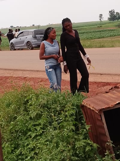 The image depicts a rural or semi-rural scene in Agwala, Nigeria. Two young Black women are walking side-by-side on a dirt road, with a light blue minivan and several other individuals visible in the background, suggesting a public area or roadside gathering. The woman on the left wears a light blue tank top and distressed denim jeans. The woman on the right is dressed in a black outfit, including a long-sleeved top and flared pants, with distinctive white ruffled cuffs. She is holding a yellow object, possibly a flower or a piece of fabric, in her right hand.

The setting includes a roadside with a paved surface and a reddish-brown dirt shoulder. Beyond the road, a vast green field, likely agricultural land, stretches towards a hazy horizon dotted with trees and scattered buildings, indicating a rural landscape. A section of a rusty corrugated metal structure, possibly part of a small shelter or debris, is visible in the foreground on the right. The sky is overcast and the lighting suggests it is daytime, perhaps late afternoon, with diffused light. The overall mood appears casual and perhaps observational, with the women seemingly engaged in a conversation or simply walking together. There is no visible text in the image.