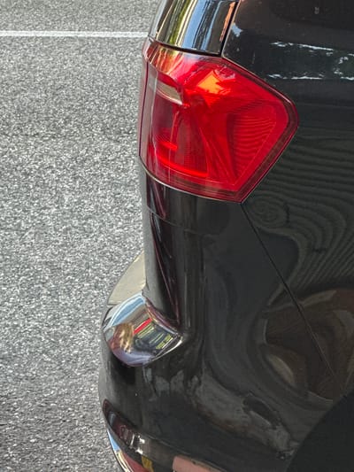 The image is a close-up, exterior shot of the rear portion of a dark-colored vehicle, likely a car, parked on asphalt with a white line visible in the upper left quadrant. The vehicle's red taillight is prominently displayed, featuring a faceted design with internal reflective elements. A portion of the car's bumper, adorned with chrome trim and reflecting the surrounding environment, is visible in the lower part of the frame. The background consists of textured asphalt, characteristic of a street or parking area. The scene is illuminated by natural light, suggesting an outdoor setting, possibly in Trieste, Italy, given the context. There are no people or discernible actions depicted in the image.