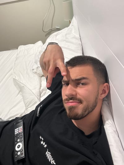 A man with short dark hair and a beard is lying in a bed covered in white sheets, possibly in a hotel room in Hampton, United States of America. He is wearing a black t-shirt and holding a TV remote. He is making a hand gesture, possibly a peace sign. The background includes a wall and a power outlet.