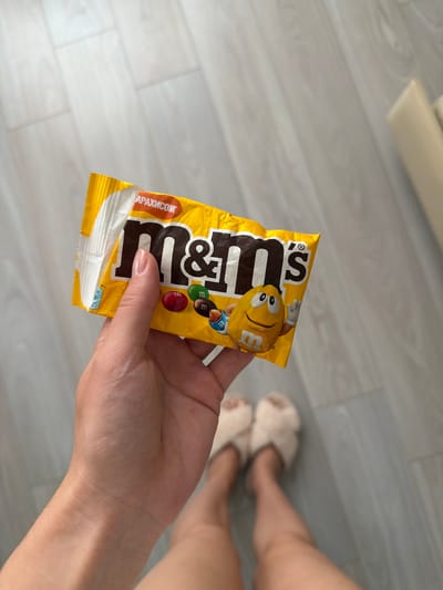 A person's hand is holding a yellow M&M's candy package. The package has the M&M's logo and images of M&M's candies. The person is wearing fluffy slippers and their legs are visible in the background. The scene appears to be indoors, on a light-colored wooden floor. This image was taken in Semiluki, Russia.