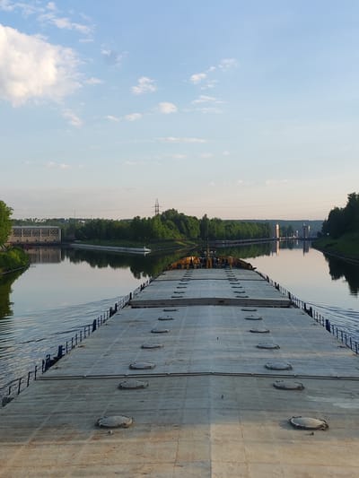 View from the deck of a barge traveling on a canal in посёлок опытного хозяйства «Ермолино», Russia. The barge deck is flat and grey with dark round hatches. The water is calm and reflects the sky and the green trees lining the canal banks. In the distance are structures, including what appear to be towers and a building. Power lines stretch across the scene above the trees. The sky is blue with scattered white clouds.
