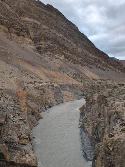 The image depicts a stark, mountainous landscape with a river flowing through a gorge. The terrain is characterized by rugged, stratified rock formations in shades of brown and tan, with scree slopes leading up to a heavily clouded sky. The river is wide and appears to be carrying sediment, giving it a murky, grayish-brown color. The banks of the gorge are steep and rocky, with some sparse vegetation clinging to the upper slopes. The overall impression is one of a remote and somewhat desolate environment. The image does not contain any individuals, actions, or interactions. The location provided, Dhar Sajam, India, is a city; however, the scene appears to be a natural geographical feature, possibly a river valley within a mountainous region of India, rather than an urban setting.