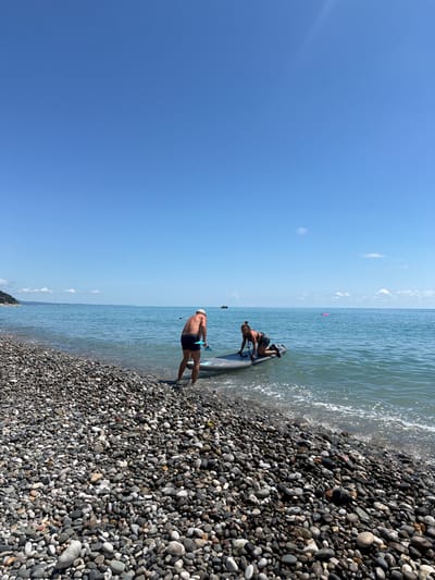 In Tsandrypsh, Abkhazia, a clear, blue sky stretches over a tranquil sea. A pebble beach lines the shore, where two individuals are engaged with a paddleboard. A man, wearing dark swim trunks and a light-colored cap, stands at the edge of the water, holding a paddle. Beside him, a woman kneels on the paddleboard, which is partially submerged in the gentle waves. The paddleboard is gray with black accents. In the background, the calm sea extends to the horizon, with a distant boat visible. The overall scene depicts a leisurely day by the sea.