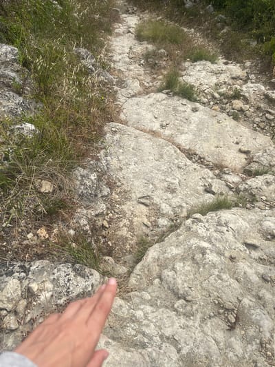 The image depicts a rough, rocky path winding through a dry, grassy hillside. Large, weathered gray rocks dominate the foreground and midground, interspersed with sparse tufts of dry grass and some green vegetation. A human hand, appearing to be reaching out, is visible in the lower left corner of the frame, its index finger pointing towards the path ahead. The path itself is uneven, made of smaller stones and dirt, and continues uphill into the distance, where it disappears into more vegetation. The overall impression is of an arid, outdoor environment, likely a hiking trail in a rural or semi-rural area. Given the information about the location, this scene is situated within the municipality of Sintra, specifically the parish of São João das Lampas, Portugal.