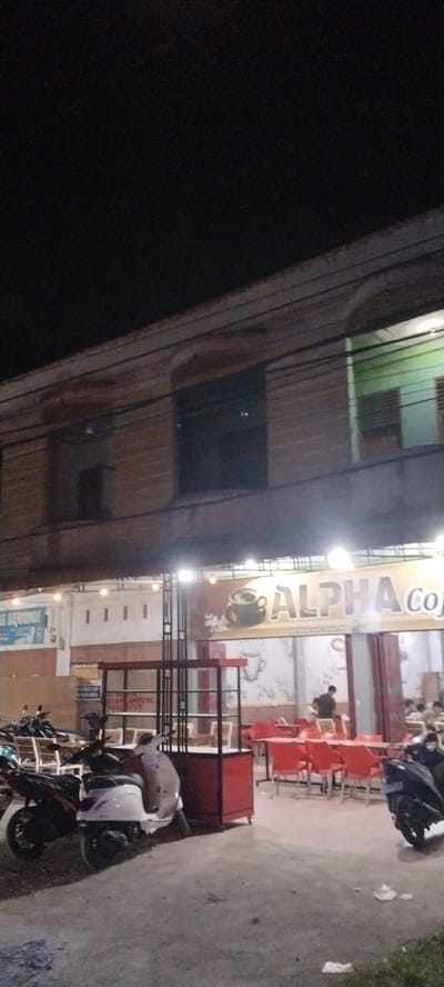 The image captures a nighttime street scene in front of a building with multiple floors. The building appears to be made of brick and has dark windows. A sign on the second floor reads "ALPHA Coffe" in large letters, accompanied by an image of a coffee cup. The ground floor is open, revealing a seating area with red chairs and tables. Several motorcycles are parked in front of the building. The ground is paved with concrete, and the overall lighting suggests it is evening or night. The presence of the coffee shop sign and motorcycles indicates it's a commercial area.