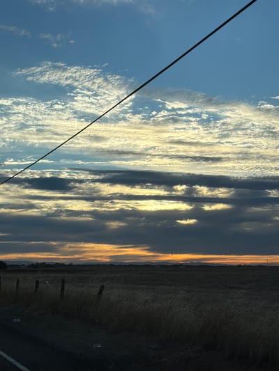 A wide shot depicts a vast, flat field with dry, tall grass. A wire stretches diagonally across the upper half of the frame, dividing the image into sky and land. The sky is a mix of blue and white, with layered clouds. The lower portion of the sky, near the horizon, is orange and yellow, suggesting either sunrise or sunset. A thin, dark line of trees and distant structures are visible on the horizon. A dirt road runs along the left edge of the frame, with several fence posts and some scattered debris on the ground. The scene is devoid of any people or animals.