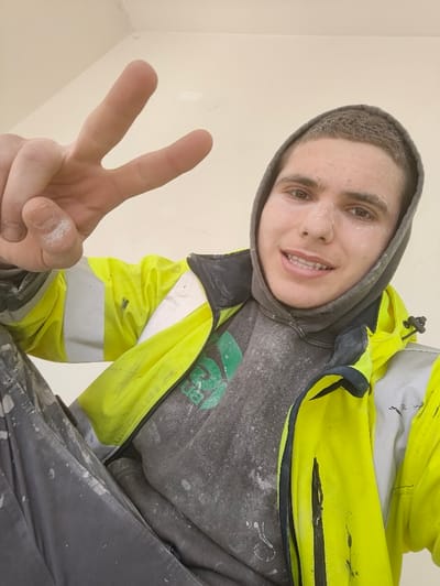 The image displays a male individual, appearing in his late teens or early twenties, posing for a photograph indoors in Deerlijk, Belgium. He has short, dark hair and wears a grey hooded sweatshirt with the hood pulled up, covering his head. A bright neon yellow high-visibility jacket with grey reflective strips is worn over the sweatshirt. Both the sweatshirt, which has a partially visible green logo on the left chest, and the jacket are heavily soiled with white particulate matter, consistent with dust or construction material. Dark grey trousers, also coated in white particulate, are visible in the lower left. The individual's face shows light white particulate matter and a wide smile, revealing upper teeth that may include orthodontic braces. His left hand is raised, forming a two-finger "peace" gesture, and is also covered in white particulate. The background consists of a plain, light-colored wall or ceiling, appearing to be an unfinished interior space. The scene is consistent with a construction or renovation environment.