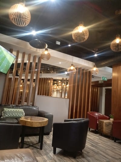 A warm and inviting lounge area is depicted, likely within a cafe, hotel lobby, or public waiting space in Pretoria, South Africa. The scene features comfortable seating and warm, atmospheric lighting, suggesting an evening or perpetually dim indoor setting.
The space is furnished with dark leather sofas and armchairs, including several in dark brown/black and two in red. One dark sofa is adorned with a green and white geometric pattern cushion. A unique side table with a live-edge wooden top and black metal legs stands between two dark seats. Another table with a distinctive, patterned wooden top is positioned near the red armchairs.
Numerous pendant lights with intricately designed wooden, slatted shades hang from the dark ceiling, casting a warm glow and creating noticeable lens flares. Vertical wooden slats serve as both decorative elements and partial room dividers, adding texture and a modern aesthetic.
On the left side of the image, a flat-screen television displays what appears to be a football (soccer) match, with visible score graphics such as "0:0" and team names, potentially including "KAIZER CHIEFS" and "SUNDOWNS" or "AMAKHOSI." In the background, a green "EXIT" sign with directional arrows is visible, indicating emergency exits within the public venue.
The overall ambiance is one of relaxation and comfort, designed for socializing or enjoying a sporting event. No people are visible in the image.