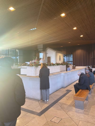The image displays the interior of a modern religious building, specifically identified as the Chapel of the Apparitions within the Sanctuary of Fátima, located in Fátima, Portugal.
The central feature is a white, gabled chapel-like structure, reminiscent of the original Chapel of the Apparitions, positioned behind a low, light-colored concrete barrier. This barrier is extensively decorated with vibrant floral arrangements in red, orange, yellow, and green. Inside the chapel structure, a white statue of the Virgin Mary is visible, and a crucifix is mounted on its exterior. Further to the right on the chapel's exterior wall, a blue and white depiction of the Virgin Mary is present.
The building's interior features a modern design with a light-colored wooden slatted ceiling containing square recessed lighting fixtures. Large windows on the left side admit significant natural light, creating a prominent lens flare in the foreground. The floor consists of light-toned tiles arranged in geometric patterns, with linear metal grates embedded. Dark brown drapes cover a wall section in the background to the right.
Approximately six individuals are present. In the mid-ground, a woman with reddish-brown hair, wearing a dark long coat and light grey wide-leg trousers with white sneakers, stands facing the central chapel, seemingly in prayer or contemplation. To her left, partially obscured by the lens flare, another person wearing a dark jacket is visible from the back. On the right side of the image, two men are seated on a wooden bench with metal supports: an older man with grey hair in a dark suit is leaning forward, looking down, while another man in a grey puffer vest sits beside him. Two other individuals are partially visible behind the concrete barrier in the mid-ground. No direct interactions between individuals are observed; all appear to be in quiet reverence.
