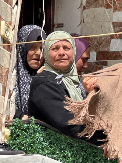 Three women are observed from an elevated perspective in Dura, Palestinian Territory, looking out at an unseen event. The central figure, a middle-aged woman, wears a light olive-green hijab and a dark long-sleeved top. Her eyes are red-rimmed and appear moist, and her mouth is slightly ajar, suggesting distress. A small white clip is fastened to her top near her right shoulder. Her right hand holds a corner of a thick, light brown, fringed woven fabric. To her left and slightly behind, an older woman with visible facial wrinkles wears a black hijab patterned with white elements; her expression is similarly distressed, and her right hand rests lightly on the central woman's left shoulder. To the central woman's right and rear, a younger woman in a purple hijab is partially visible in profile, facing towards the right with a downward gaze; her right hand also grips the fringed fabric. Below the women, a band of dense green artificial foliage is present. The background consists of a wall constructed from light tan and reddish-brown rectangular tiles in a staggered pattern. A tan corrugated conduit runs vertically on the left side of the wall, alongside a small, rectangular, light-colored electrical box. Two thin, dark horizontal wires traverse the scene across the mid-ground. A single thin, white twisted cord hangs vertically in the upper left quadrant of the frame. The scene is illuminated by bright daylight.