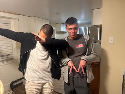 Two young males are standing in what appears to be a kitchen. The male on the left is wearing a black zip-up jacket over a gray t-shirt and light-colored shorts. He is performing a "dab" gesture with his arms. The male on the right is wearing a gray t-shirt with an "ACME" logo and dark gray sweatpants. He is holding his hands up in a peace sign gesture. The kitchen has white cabinets, stainless steel appliances, and wooden flooring. A light switch is visible on a tan-colored wall. The overall environment suggests an indoor, residential setting, possibly a kitchen or dining area. No specific city or state is identifiable from the visual information provided.