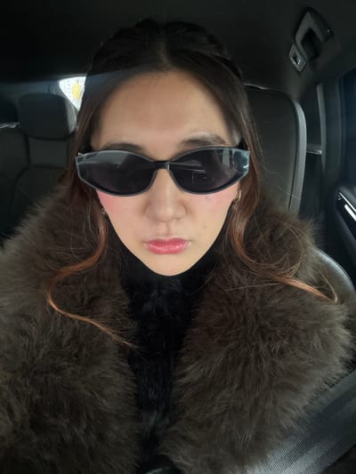 A woman wearing sunglasses poses indoors. She has dark hair, with some strands a lighter reddish-brown color. She wears dark clothing with a brown fur-like collar. The picture appears to be taken inside a car, with parts of the car interior visible in the background.