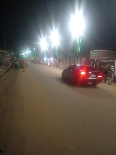 The image depicts a street scene at night in Tundun Wada West, Nigeria, illuminated by streetlights and the taillights of a dark-colored car. The road surface is paved and appears to be made of asphalt or a similar material. The car, a sedan with its brake lights illuminated, is in motion on the right side of the road. Along the sides of the street, there are buildings and structures, some of which appear to be shops or homes. Several people are visible, including individuals standing on the side of the road and a person standing near the car. There are also motorcycles parked along the side of the road. The overall impression is that of a moderately busy street at night, with artificial lighting providing visibility.