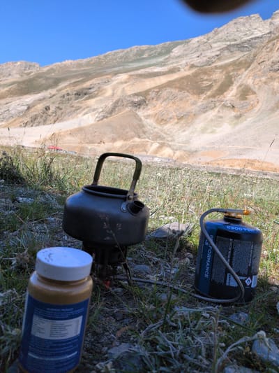 The image captures an outdoor scene with a mountainous, arid landscape in the background under a clear blue sky. In the foreground, on uneven ground covered with dry grass and rocks, are camping equipment. A dark metal camping kettle sits on a stove. To its right is a blue and black gas canister with a grey hose. In front of the kettle, a jar with a white lid is visible, with a label partially obscured. A small red building is faintly visible in the middle ground of the landscape. The location is identified as Matayeen, India. There are no people present in the image.