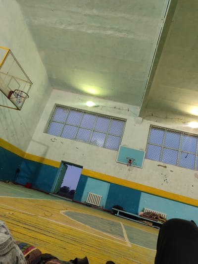 This media file depicts the interior of a dimly lit gymnasium, likely located in Boroldoy, Kyrgyzstan. The scene is dominated by a well-worn wooden basketball court, marked with faded lines in yellow and turquoise. The walls are painted in alternating bands of blue and yellow, showing signs of age and wear.
Two basketball hoops are visible: one larger hoop with a backboard hangs high on the left wall, while a smaller, child-sized hoop is mounted lower on the right wall. Large windows with metal grates are set high on the walls, allowing a glimpse of the dusky sky outside, suggesting it is either early morning or late evening. Fluorescent lights, some appearing to be malfunctioning, illuminate the space unevenly.
On the right side of the gym, a long bench holds several rolled-up blankets or sleeping mats, and a backpack. A radiator is mounted on the wall above the bench. In the foreground, the edge of a person's leg, covered in dark fabric, is visible, along with the patterned fabric of what appears to be a blanket or cushion, implying someone is resting or sleeping on the floor. Another person's patterned clothing is visible on the left side of the frame.
A red bucket is placed near an open doorway on the left, through which a dimly lit outdoor scene with trees can be seen. The overall atmosphere is one of quiet solitude and perhaps a sense of makeshift living arrangements within the gymnasium. There is no visible text in the media file.