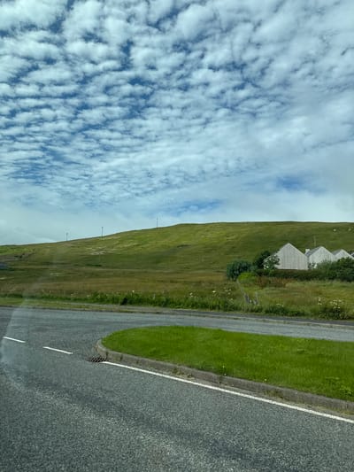An exterior, eye-level shot captures a cloudy sky and a green, grassy hillside in Aithsetter, United Kingdom. The foreground features an asphalt road with white lane markings and a concrete curb bordering a strip of lush green grass. In the middle ground, the hillside is dotted with sparse vegetation and several tall, thin poles that appear to be streetlights or wind turbines. At the base of the hill, a cluster of pale-colored houses with distinctive gabled roofs is nestled amongst trees and shrubs. The overall impression is one of a rural landscape under a partly cloudy sky.