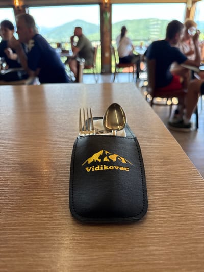 The media file shows a close-up of cutlery resting in a black leather pouch on a wooden table. The pouch has a golden embroidered logo featuring a mountain and the text "Vidikovac". In the background, through a large window, a blurred scene of people sitting at tables and a mountainous landscape is visible. The lighting suggests it is daytime. The overall scene appears to be a restaurant or cafe setting, possibly in Miločani, Montenegro, given the location context.