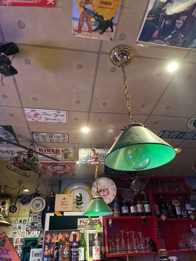 The image depicts the interior of what appears to be a bar or restaurant with a vintage, eclectic theme, decorated with various signs, memorabilia, and a chandelier. The ceiling is made of textured panels and is illuminated by spotlights and the central chandelier.
Prominently featured is a hanging green glass lampshade with a visible lightbulb. Various signs are affixed to the ceiling, including one for "New York Yankees," a "Historic US 66" sign, and a "Biker Parking Only" sign with a cartoon character. Other decorative elements include a miniature helicopter, a motorcycle, and several framed pictures or posters.
On a red shelf in the background, an assortment of bottles, likely alcoholic beverages, are displayed. Glasses are also visible on the shelves. There are also clocks and a box labeled "Don Julio 1942."
The overall impression is a cluttered yet deliberately curated space, filled with items evoking American nostalgia and popular culture. There is no visible indication of the location being in Adachi, Japan.