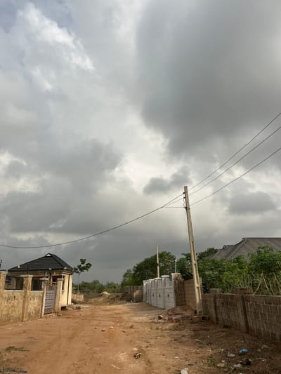 This image depicts a semi-developed area within Ilishan, Nigeria, under a heavily overcast sky. A reddish-brown dirt road, showing signs of vehicle tracks, occupies the foreground and extends into the midground. Litter, including plastic bags, is visible on the roadside.
To the left, a light-colored building with a dark, sloping roof is partially enclosed by a low, plastered wall with a light-colored metal gate. A single green tree stands behind this structure.
In the midground, towards the center-right, a concrete utility pole supports multiple overhead electrical wires that traverse the scene horizontally. Adjacent to the pole and extending to the right is a grey block perimeter wall, interspersed with newer, lighter grey pre-cast concrete fence panels. Beyond these walls, additional buildings with dark roofs are partially obscured by green vegetation, including small trees and shrubs. The background reveals undeveloped land with sparse greenery under the distant, hazy sky. No individuals are present in the scene.