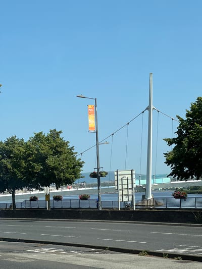 The image depicts a street scene in the city of Derry/Londonderry, United Kingdom, with the Peace Bridge prominently visible in the background. The foreground shows a paved road with white markings, suggesting a street or highway. Adjacent to the road is a low, dark stone wall topped with a black metal fence. Along the fence, several large planters filled with red and pink flowers are strategically placed.

Behind the fence, a wide body of water, likely the River Foyle, stretches across the middle ground. The iconic Peace Bridge, a pedestrian and cycle bridge, spans the river. Its distinctive white structure, with suspended cables, is clearly visible against the clear blue sky.

On the left side of the image, lush green trees line the embankment. A lamppost stands on the sidewalk, adorned with an orange and yellow banner. The banner appears to have some text and graphic elements, though they are not clearly decipherable from this distance. Further along the street, another lamppost can be seen.

On the right side of the image, another large green tree partially obscures the view of the bridge. The sky is a vibrant, cloudless blue, indicating a sunny day. The overall scene captures a typical urban riverside setting, highlighting a key landmark of Derry/Londonderry.