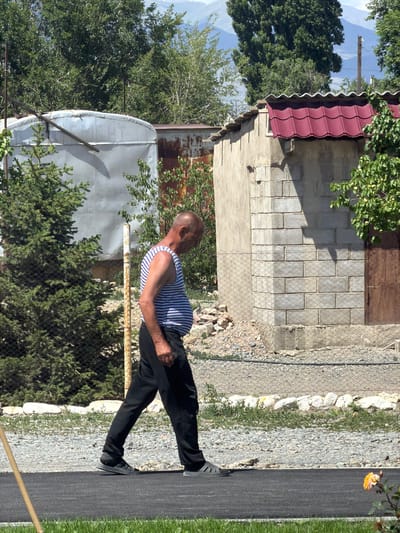 A man with a shaved head and a striped tank top stands on a paved path. He is wearing black pants and black shoes. He is facing away from the camera, looking to his left. Behind him, there is a fence, some trees, and a building with a red roof. The scene appears to be outdoors in a rural or semi-rural setting. The lighting suggests it is daytime, likely morning or afternoon. The image's orientation is vertical, with the man positioned on the right side of the frame. There are no visible text elements in the image.