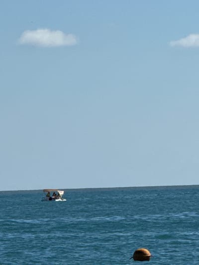 In the city of Adlerskiy, Russia, a wide shot captures a calm, blue sea under a pale blue sky with two small, white clouds. In the distance, a small boat with a canopy carries several people. In the foreground, a brown buoy floats on the water. The horizon is clearly defined, separating the sea and the sky.