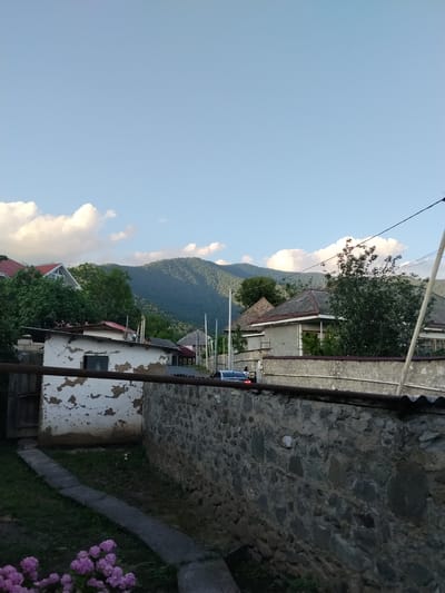 A rural village scene is depicted under a clear blue sky with scattered white clouds. In the foreground, a weathered stone wall runs horizontally across the image, topped with a rusted metal pipe. To the left of the wall, a small, dilapidated white building with peeling paint and a single window is visible. A narrow, concrete path leads from the building into the foreground, where a cluster of vibrant pink flowers is in bloom.

Behind the foreground wall, several houses with pitched roofs can be seen, nestled amongst lush green trees. The architecture suggests a traditional style, with some houses featuring grey stone or stucco exteriors. In the background, rolling, tree-covered mountains dominate the landscape, their slopes softened by the distance and hazy light. A few power lines are strung across the top of the scene.

A dark-colored car is parked on a road behind the stone wall, with two figures near it. The lighting suggests it is late afternoon or early evening, with the sun casting a warm glow over the scene, highlighting the texture of the stone wall and the vibrant colors of the flowers. The overall atmosphere is peaceful and serene, characteristic of a quiet village setting. The location is identified as Oghuz, Azerbaijan.