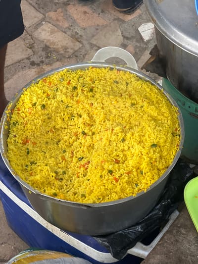 A large metal pot overflowing with bright yellow rice is centrally positioned in the image. The rice is mixed with small, diced pieces of red and green vegetables, likely peppers and scallions. The pot has two metal handles and appears to be sitting on a blue cooler. To the right of the pot, a large silver pot with a dark lid is visible. The ground beneath the pot is paved with irregular, earthy-toned stones, some of which appear wet. In the upper left corner, a person's dark-colored shorts and part of their leg are visible, suggesting they are standing near the pot. The overall scene appears to be an outdoor setting, possibly a market or a street food stall in Ijegun, Nigeria, given the context.