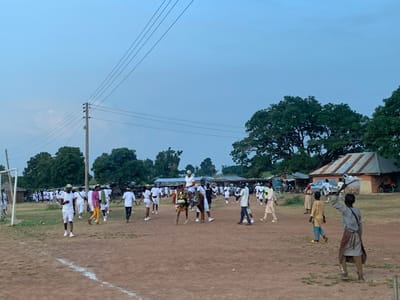 The image depicts an outdoor scene in Kubi A, Nigeria, characterized by an open, unpaved field under a light-blue, overcast sky. Numerous individuals are present, predominantly young adults dressed in matching white shirts and trousers, with several wearing light green or white caps/helmets. This group is spread across the central and left portions of the field, generally moving from left to right and towards the background.
In the center foreground, a person in a detailed, multi-colored costume with gold, black, and possibly red elements is visible, moving among the white-clad individuals. Further back, another person in white is being carried on someone else's shoulders. On the far left, a makeshift goalpost with a net is visible. A white line is painted on the brown earth in the lower-left foreground.
On the right side of the frame and in the background, several single-story structures with corrugated metal roofs are visible, interspersed with large, dense green trees. A light-colored car and partially visible motorcycles are parked near these buildings. Utility poles with multiple overhead power lines traverse the upper-left quadrant of the image. In the right foreground, a person in light brown clothing, facing away from the camera, holds a long stick with a white cloth or flag attached, extended upwards and to the right. Other individuals in various attire are scattered on the field and near the structures, observing or moving.