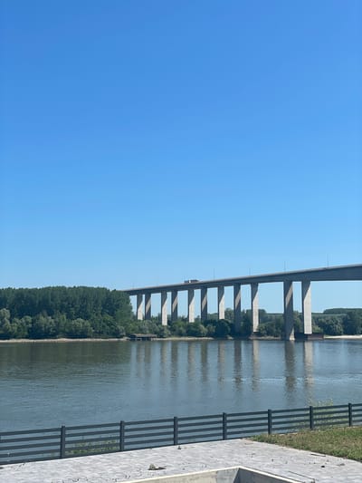 The image shows a long bridge over a body of water, possibly a river, in Indjija Municipality, Serbia. The bridge is supported by a series of concrete pillars that cast shadows on the water. A line of trees and vegetation is visible on the riverbank beneath the bridge. The sky is clear and blue. There is a fenced walkway on the right side of the image. A small vehicle is visible on the bridge.