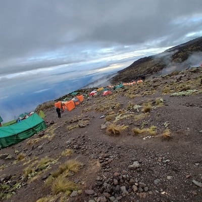 The image depicts a high-altitude mountain encampment at Gilman's Point, Tanzania, situated on a dark, rocky, and sparsely vegetated slope. Numerous tents are visible across the uneven terrain; large green tents occupy the foreground-left, while a prominent line of orange and blue tents stretches across the mid-ground. Further up the slope, a denser cluster of multi-colored tents (green, orange, red, blue) recedes into a misty background. One individual in dark clothing stands in the mid-ground between two orange tents. The sky above is heavily overcast with grey clouds. A vast layer of clouds forms a distant horizon below the camp, indicating the high elevation of the site. Mist partially obscures the higher mountain slopes to the right.