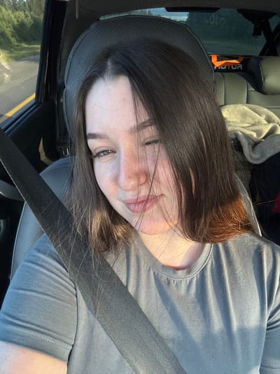 A young Caucasian woman with long, dark brown hair and fair skin is seated in the passenger seat of a car, wearing a gray t-shirt and a black seatbelt. She is looking towards the camera with a slight smile and one eye partially closed. The interior of the car is visible, including the headrest, car seat, and the window. Outside the car, a blurred background suggests a road and trees, indicating the car is in motion. The lighting suggests it is daytime, with sunlight casting shadows across the woman and the car's interior. The setting appears to be a rural or suburban road, possibly in Krasnokamsk, Russia, given the prompt's context.