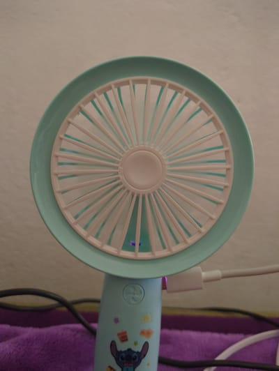 A teal and white portable fan is displayed prominently in the foreground against a neutral, textured background. The fan features a circular grille with numerous white spokes radiating from a central hub. A light purple glow emanates from beneath the fan blades. The base of the fan is also teal and adorned with colorful cartoon decals, including a character resembling Stitch from Disney's Lilo & Stitch. A white USB charging cable is plugged into the side of the fan's base. To the left of the fan, a dark grey cable is visible, resting on a soft, purple fabric surface. The scene is static and does not depict any action or interaction. The location is identified as Rusiec, Poland.