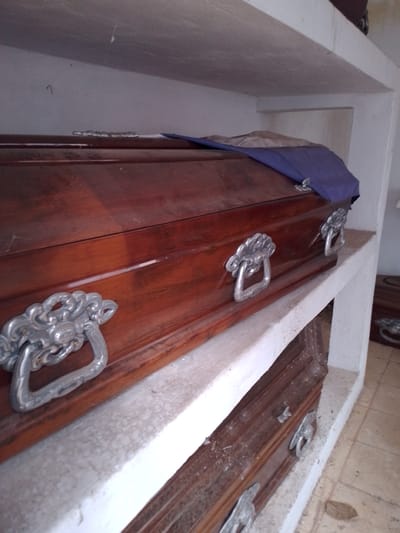 The image depicts two wooden coffins stored in a white concrete niche within what appears to be a mausoleum or cemetery. The top coffin is varnished and has ornate silver-colored handles, and is partially covered by a blue cloth. The lower coffin, also with silver-colored handles, appears dustier. The location is inferred to be in Asunción, Paraguay. The image suggests a place for the deceased with stacked burials.