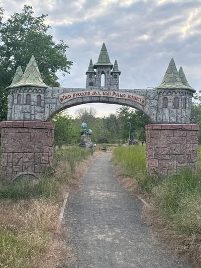 An ornate archway resembling a castle gate stands prominently in a park-like setting, welcoming visitors down a gravel path. The arch is constructed of gray, faux stone blocks and features conical turrets topped with green, shingle-like roofs. Inscribed in red Cyrillic lettering on the arch reads, "Там русский дух, там Русью пахнет" which translates to "There is Russian spirit, there it smells of Rus'". Flanking the arch are cylindrical pillars, also made of faux stone, with a rough, reddish-brown texture. The path leads towards the arch, disappearing into the distance. Lush green trees and foliage surround the area, with dry, tall grass growing along the sides of the path. In the background, a sculptural element resembling a blue orb atop a rocky base is visible. The sky above is overcast with some patches of sunlight breaking through. This archway is a recognizable landmark in the city of Yeysk, Russia, specifically located in the Victory Park (Park Pobedy).