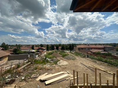The image shows an outdoor construction site in Dolni Dabnik, Bulgaria. The site is partially built, with piles of dirt, gravel, and lumber visible. A partially constructed wall is being plastered by a worker in a white shirt and yellow pants. The background features a residential area with numerous houses with red tile roofs, surrounded by trees. The sky is partly cloudy.