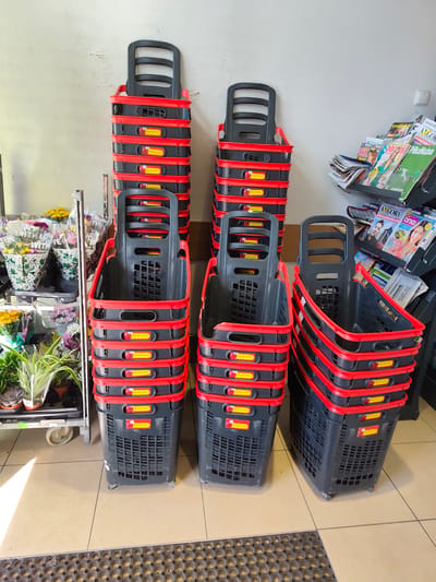 This image depicts a retail environment, likely a grocery store, in Łagiewniki Nowe, Poland. The foreground features stacks of black plastic shopping baskets with red trim, arranged in neat columns against a light-colored wall. Each basket has a prominent yellow label with a red ladybug logo and the text "Biedronka," indicating the store's brand. To the left, a display rack holds various potted plants and bouquets of flowers, some wrapped in plastic. In the background, to the right, a magazine rack is filled with a selection of publications. The floor is tiled, and a textured mat is visible at the bottom of the frame. The lighting suggests an indoor setting with natural light entering from the left. There are no people visible in the image.