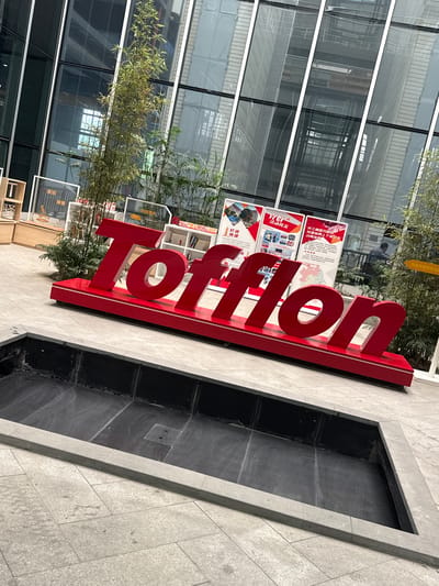 An exterior shot captures a large, red, three-dimensional "Tofflon" logo positioned on a flat, light gray tiled surface. The logo rests on a red platform with white trim and is situated in front of a modern building with a glass facade. The building's interior, visible through the glass, appears to be an atrium or open space with multiple levels and structural elements. Behind the logo, there are various informational posters displayed on white stands, some featuring images and text. To the left and right of the logo, there are potted plants, including tall bamboo-like specimens and smaller leafy plants. A dark, rectangular indentation in the foreground, likely a dry fountain or water feature, is also visible. The scene suggests an outdoor area within a commercial or corporate setting. The city of Minhang District, China, is implied as the location.