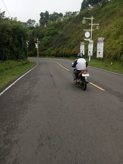 An individual is depicted riding a dark-colored motorcycle with an orange-brown seat away from the viewer along a two-lane asphalt road in the city of Deothang, Bhutan. The rider wears a white and black helmet, a white short-sleeved shirt with a black band, and gray pants. The motorcycle's rear license plate is partially visible as "N 481". The road curves leftward, featuring a solid white line marking the left edge and a solid yellow line separating the lane on the right. Cracks are visible in the asphalt. To the left of the road, a grassy verge, a low wall or curb, and dense green foliage and trees are present. To the right, a steep, heavily vegetated hillside rises, adorned with multiple decorative white poles featuring lantern-like fixtures and circular signs. Four vertical banner signs, white with text, hang beneath one of these poles. The sky is overcast and light gray.