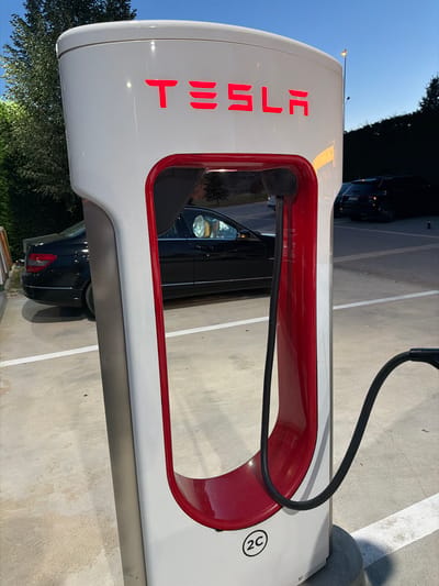 This is a portrait-oriented, close-up shot of a Tesla Supercharger station. The station is white with red accents around the charging port area. The word "TESLA" is illuminated in red on the side of the station. A black charging cable extends from the top of the station, curving downwards. A circular emblem with "2C" inside is visible on the lower right side of the station.

In the background, a black car is parked next to the charging station, and another dark-colored vehicle is visible further back. Trees with dark green foliage are present on the left side of the frame. The ground is a light-colored concrete surface with white lines indicating parking spaces. The sky is clear and blue, suggesting daytime, possibly late afternoon or early evening due to the shadows. The scene appears to be an outdoor electric vehicle charging area. There is no visible human presence. The overall impression is one of modern technology and infrastructure for electric vehicles.