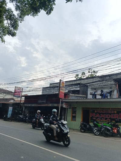 An outdoor scene shows a street with several parked motorcycles in front of storefronts. One storefront has "SAMUDERA JAYA MOTOR BB" written above its entrance. Another adjacent store reads "WARTEG MANDIRI". Two motorcyclists are driving on the street, one in front and one behind. Above the buildings, there are multiple electrical wires crisscrossing. The sky is overcast with grey clouds, and a few tree branches are visible in the top left corner. Laundry is hanging on the roof of one of the buildings.