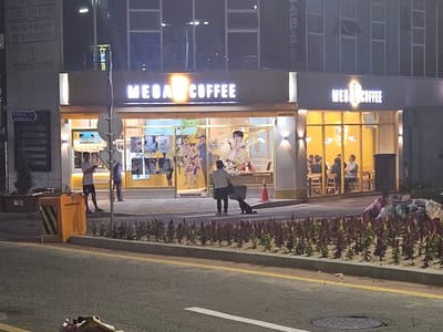At night in Eumnae-ri, South Korea, a "MEGA COFFEE" establishment is visible. The storefront features a prominent illuminated sign with the words "MEGA COFFEE" in white capital letters. Behind the large glass windows, interior lighting reveals several patrons seated at tables. To the left of the entrance, a single person stands outside, facing away from the camera, wearing a light-colored t-shirt and dark shorts. A black shopping cart is positioned next to them. A bright orange trash receptacle with diagonal black and yellow stripes is situated on the sidewalk to the left of the person. A landscaped planter box with rows of flowering plants runs along the edge of the sidewalk adjacent to the street. In the foreground, a portion of the asphalt street is visible, with yellow lane markings and a manhole cover. Several trash bags are piled up on the sidewalk near the right side of the building.