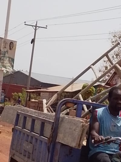 In Dungu, Ghana, a man drives a blue three-wheeled vehicle loaded with concrete blocks and metal framing. An electric pole stands in the background near a building, and a political poster is affixed to a pole on the left side of the image. Another person is visible in the background next to a brick structure.