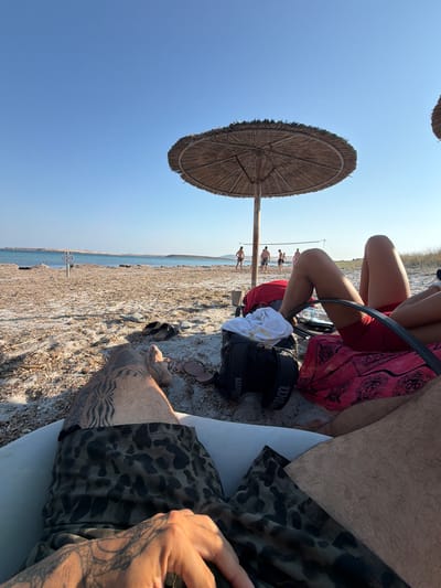 In the city of Moudros Municipal Unit, Greece, an outdoor scene unfolds on a sandy beach under a clear blue sky. The foreground is occupied by the legs of two individuals. One person, partially visible, wears camouflage-patterned shorts and has a spiderweb tattoo on their upper thigh and hand. The other person is seated, their legs extended, wearing red shorts and a pink sarong with a patterned design.
Behind them, beach chairs and bags are scattered on the sand. A straw umbrella provides shade. In the mid-ground, a volleyball net is set up, and several individuals are playing volleyball. The background features the calm sea and a distant, arid coastline. A signpost is visible near the water's edge. The overall impression is a relaxed day at the beach.